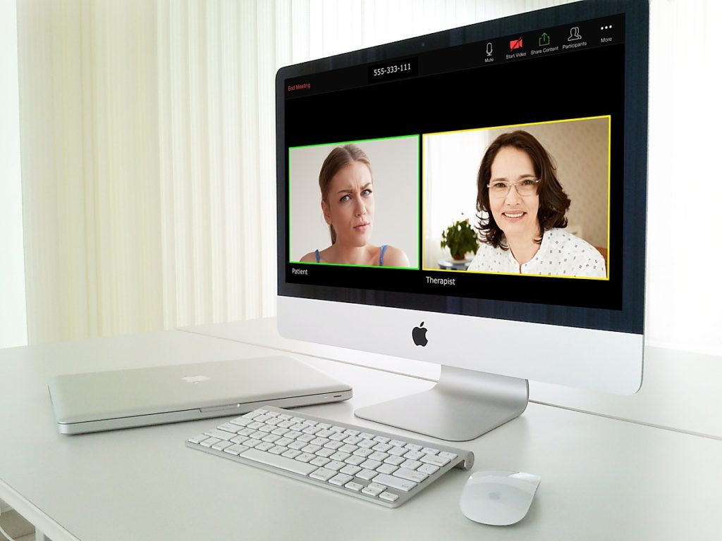 Zoom therapy sessions- Photo of a white laptop and monitor on a white desk, with white walls and blinds. On the screen is the Zoom application having a black background with leave meeting on the top left in red, a nine digit meeting number in the top center, and a series of icons for microphone, video camera, share screen, participants, and more on the top right. Centered in the screen are two display boxes side-by-side. The box on the left is outlined in bright green, labeled Patient. It shows a closeup of a young woman with her blond hair pulled back and wearing a blue top. She looks distressed. The second box is outlined in a paler color, and is labeled Therapist. It shows a slightly older woman with light-framed glasses, brown shoulder-length hair and a white top with muted dots.
